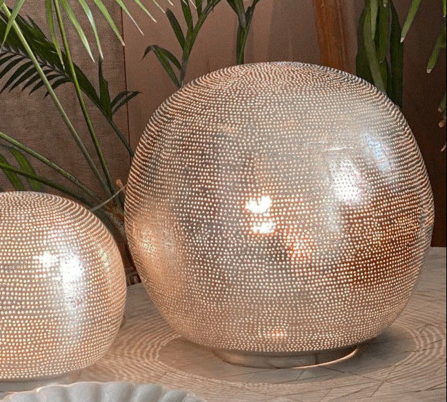 Zenza Filisky silver ball table lamps in two sizes on a marble table surrounded by tropical plants, casting warm light through punched filigree holes